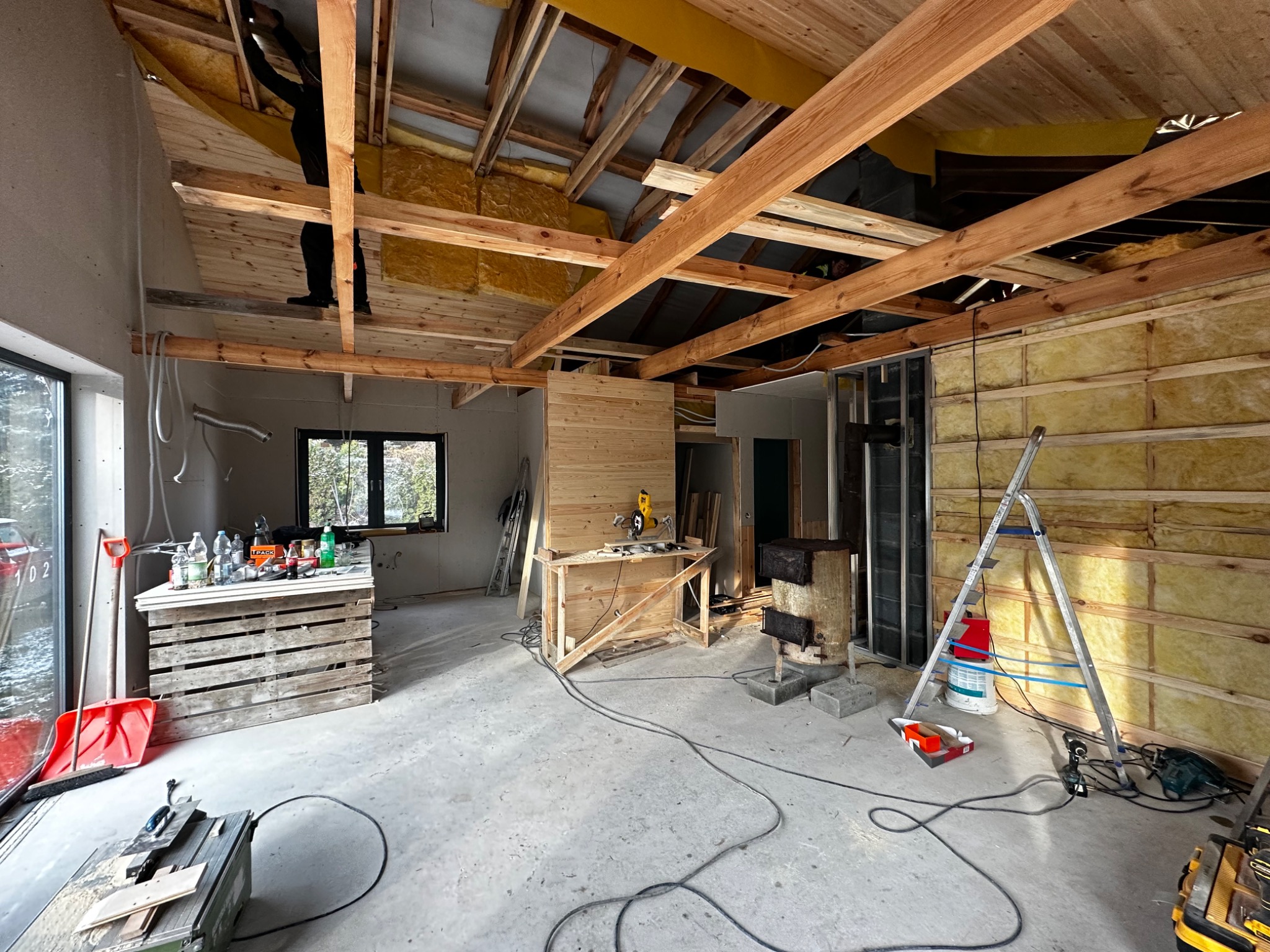 Wnętrze w trakcie remontu: widoczne drewniane elementy konstrukcyjne dachu, ocieplenie ścian wełną mineralną, prowizoryczne stanowisko pracy z narzędziami, drabina oparta o ścianę i osoba pracująca...