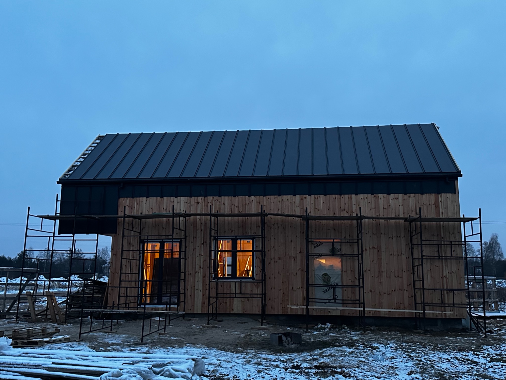 Surowy stan elewacji domu z drewnianą okładziną, widoczne rusztowania i ciemna blacha na dachu, zimowy krajobraz z resztkami śniegu na ziemi, ciepłe światło z okien