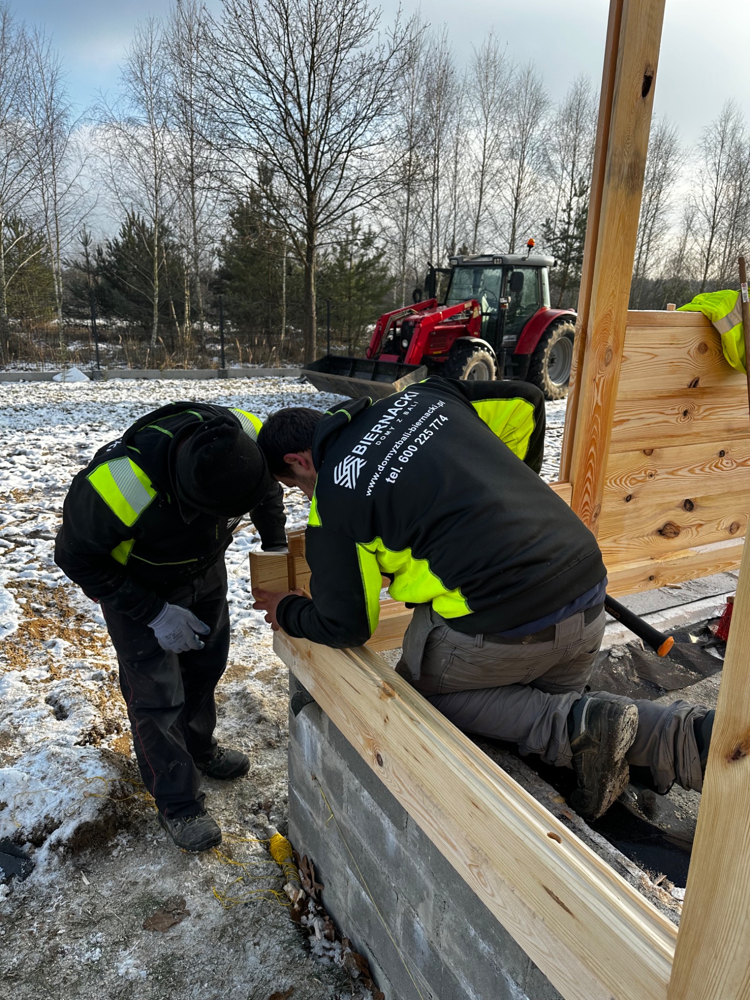Dwóch pracowników w odblaskowych kurtkach montuje drewnianą konstrukcję na fundamencie z bloczków betonowych; w tle traktor na zasypanym śniegiem terenie.
