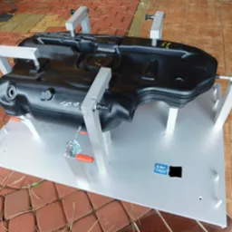 Czarny zbiornik z tworzywa sztucznego zamocowany w srebrnym oprzyrządowaniu z zaciskami, widoczne oznaczenia 'SET UP' i 'Honda D 3.255', ustawiony na metalowej płycie z logo firmy.