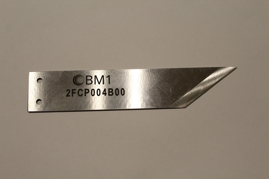 Metalowy nóż z dwoma otworami i wygrawerowanym numerem seryjnym CBM1 2FCP004B00, leżący na gładkiej powierzchni.