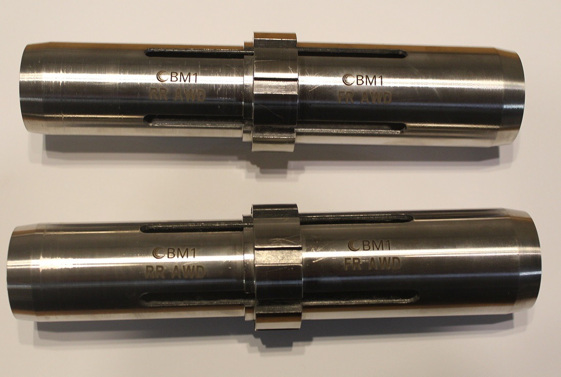 Dwa identyczne, cylindryczne elementy metalowe z wygrawerowanymi napisami 'CBM1' i 'FR-AWD' oraz 'RR-AWD', połączone pierścieniowym elementem w połowie długości, leżące równolegle na jasnym tle.