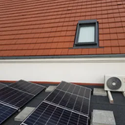 Panele fotowoltaiczne i klimatyzator zamontowane na płaskim dachu pokrytym papą, widoczne okna dachowe i część dachu z czerwonej dachówki.