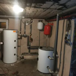 Dwa białe zasobniki ciepłej wody użytkowej w piwnicy, z widocznymi rurami instalacyjnymi, zbiornik wyrównawczy czerwony i biały, surowy betonowy sufit z lampą, ściany z widoczną strukturą, małe...