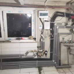 Instalacja grzewcza z rurami w izolacji, widoczne zawory i sterownik, montaż w pomieszczeniu technicznym w Rudzie Śląskiej, odbicie instalatora w szybie okna.