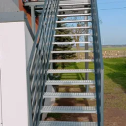 Zewnętrzne schody metalowe z ażurowymi stopniami i szarą balustradą, prowadzące do balkonu na tle zielonego pola i błękitnego nieba.