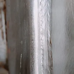 Szczegółowe zbliżenie na spoinę łączącą dwa metalowe profile, widoczna struktura spoiny. Surowy, przemysłowy wygląd.