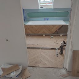 Marek Kołyt - Wnętrze łazienki w trakcie remontu: ułożona w jodełkę drewnopodobna podłoga, zabudowa wanny z płytek, przygotowane ściany z płyt kartonowo-gipsowych, widoczne narzędzia pracy i materiały budowlane.