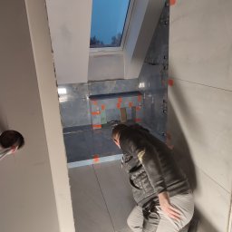 Marek Kołyt - Pracownik w trakcie układania szarych płytek w łazience z oknem dachowym, widoczne pomarańczowe elementy dystansowe i krzyżyki do fug.