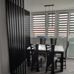 Nowoczesna jadalnia z czarnym parawanem lamelowym, białym stołem i designerskimi krzesłami pod wiszącą lampą z trzema kloszami.