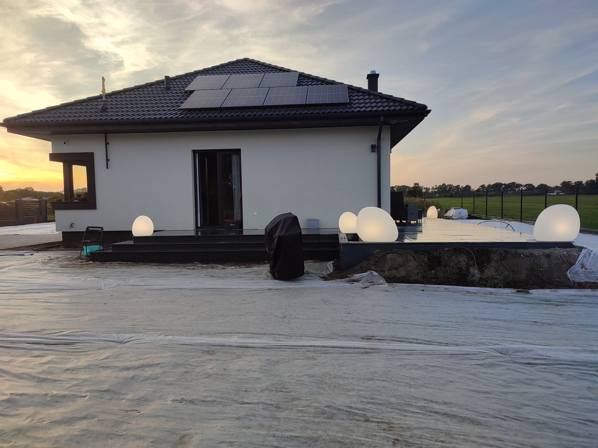 Biały dom jednorodzinny z czarnym dachem i panelami słonecznymi w trakcie remontu, folia ochronna na trawniku, oświetlenie zewnętrzne w postaci kul.