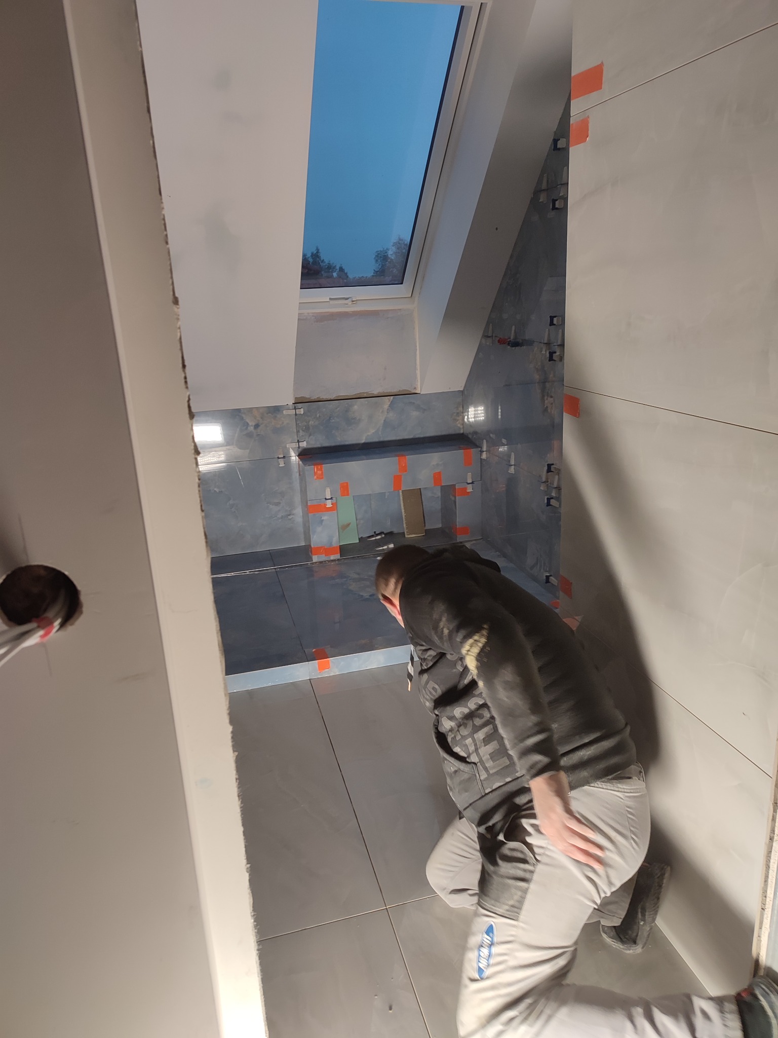 Pracownik w trakcie układania szarych płytek w łazience z oknem dachowym, widoczne pomarańczowe elementy dystansowe i krzyżyki do fug.