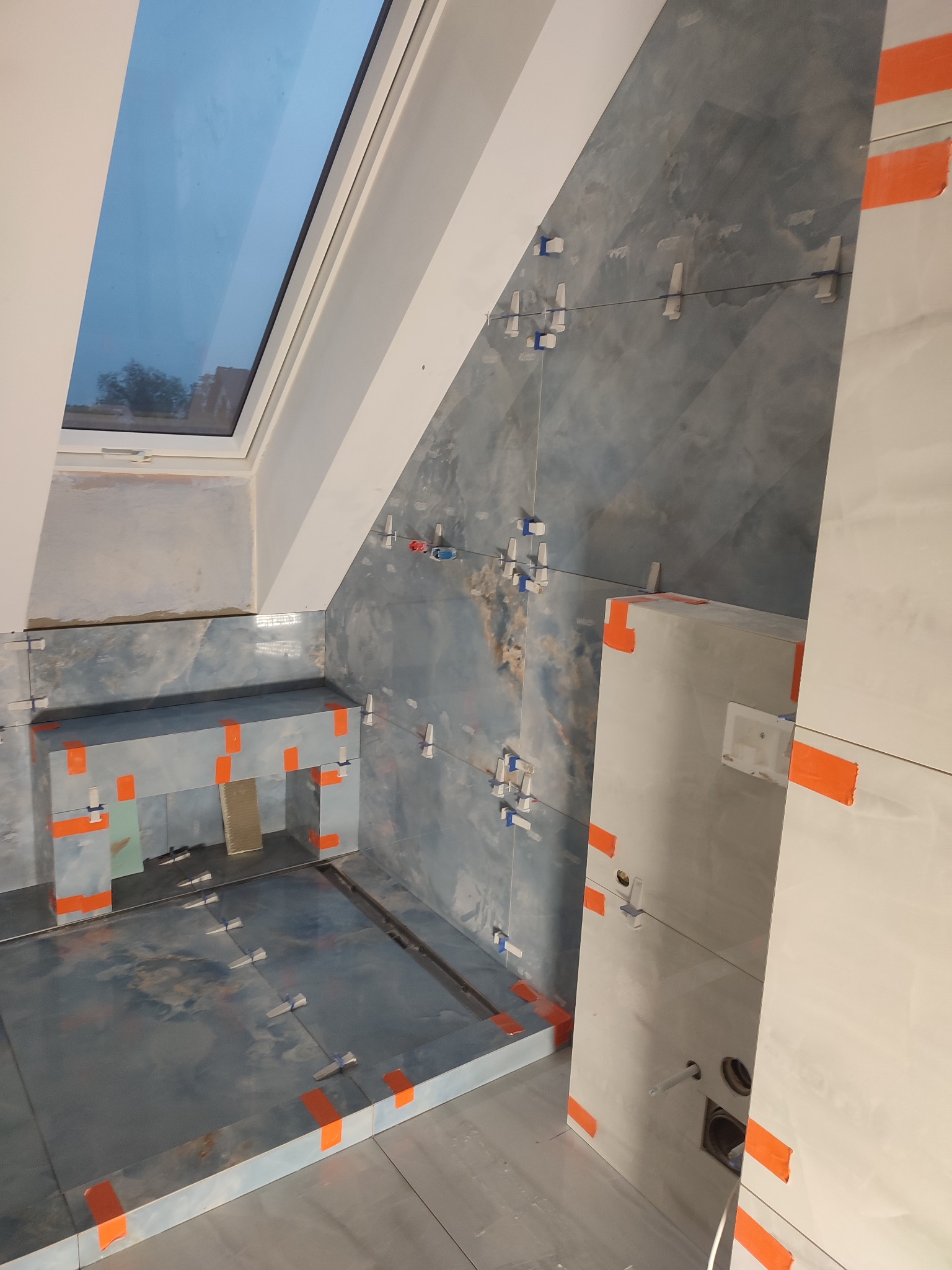 Łazienka w trakcie układania płytek: widoczne płytki w odcieniach szarości i błękitu na ścianach i podłodze, krzyżyki dystansowe, pomarańczowa taśma malarska zabezpieczająca krawędzie, okno dachowe...