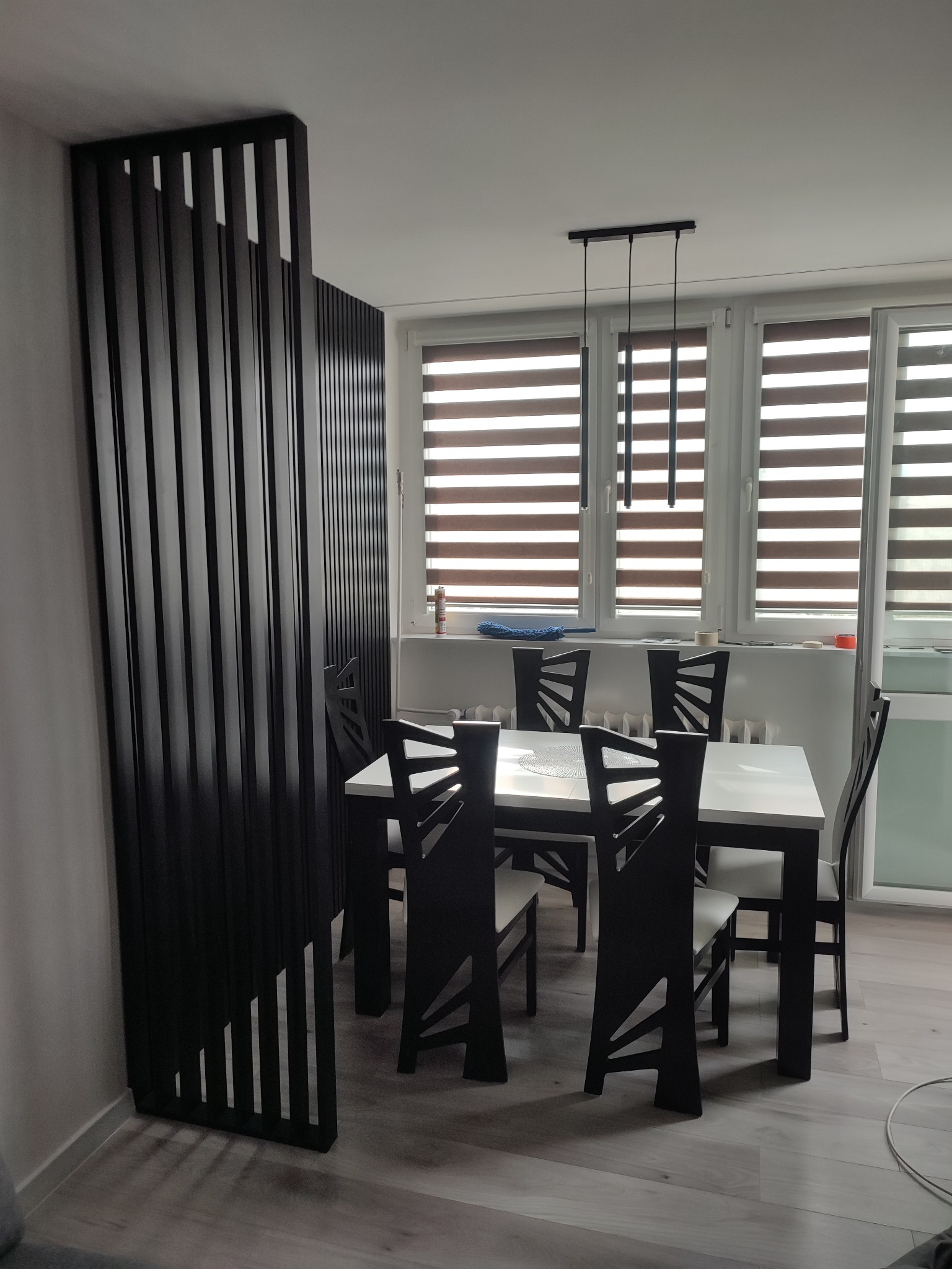 Nowoczesna jadalnia z czarnym parawanem lamelowym, białym stołem i designerskimi krzesłami pod wiszącą lampą z trzema kloszami.