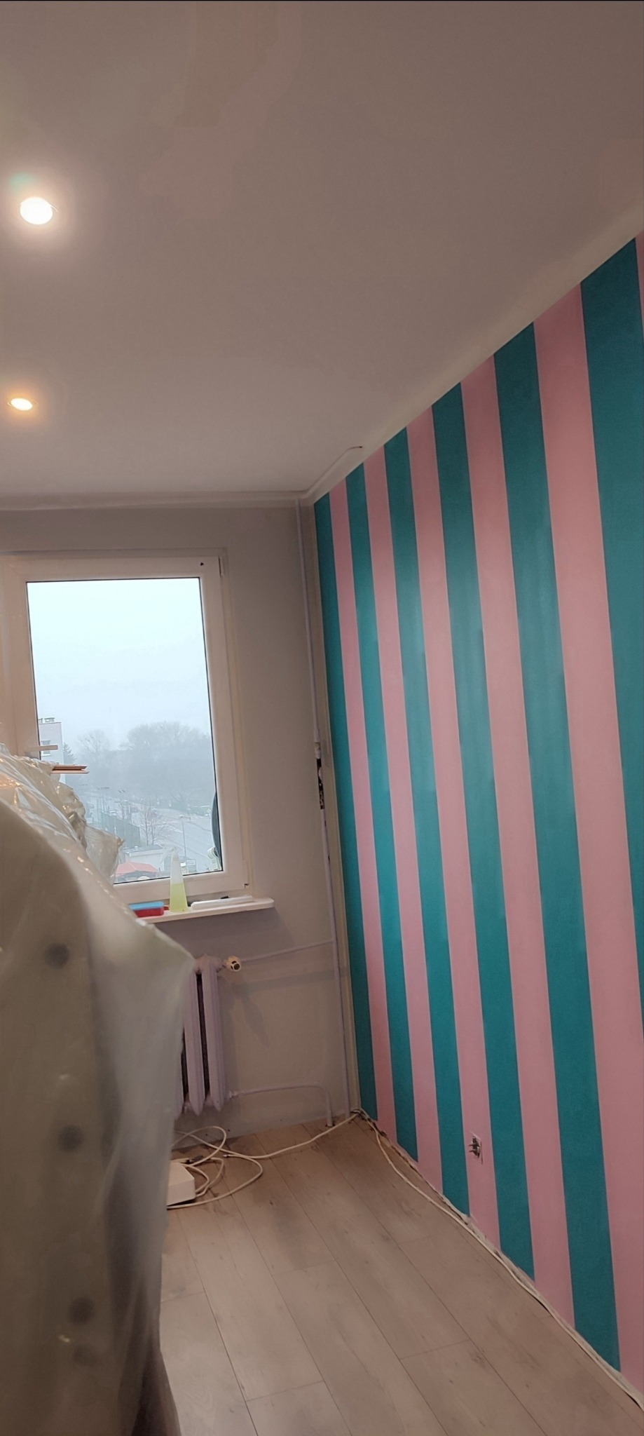 Pomieszczenie z nowo pomalowaną ścianą w pionowe pasy w kolorach różowym i turkusowym. Okno z widokiem na szarą, zimową scenerię. Część pomieszczenia zakryta folią ochronną.