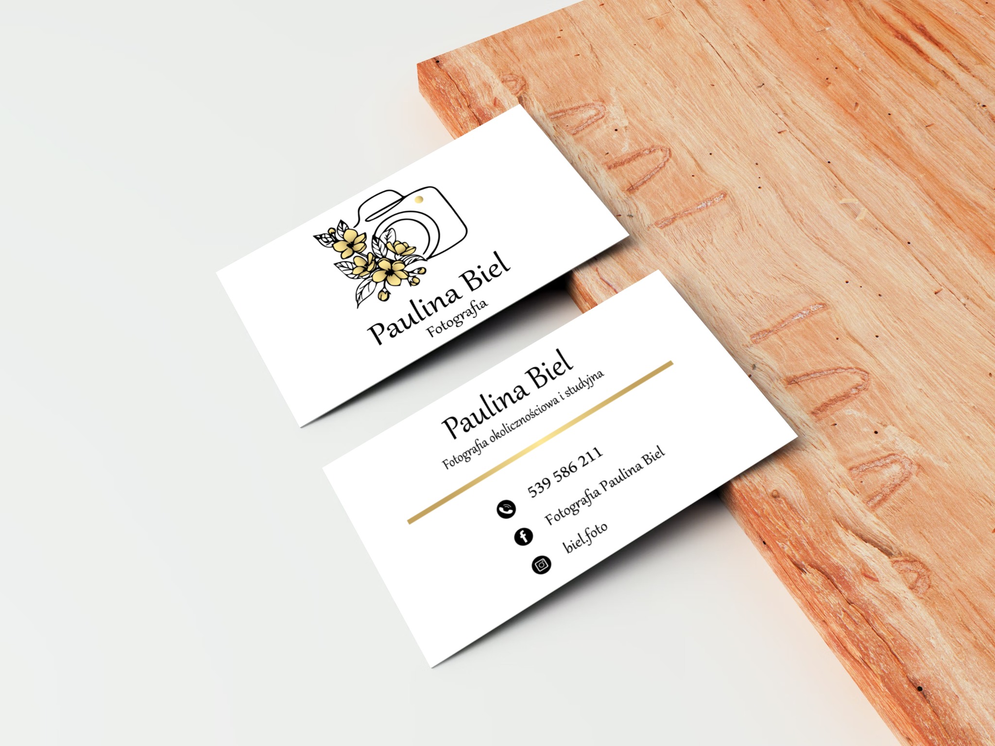 Dwie wizytówki fotografki Pauliny Biel leżą na drewnianym blacie. Wizytówki są białe, z czarnym tekstem i grafiką aparatu z kwiatami w kolorze żółtym.