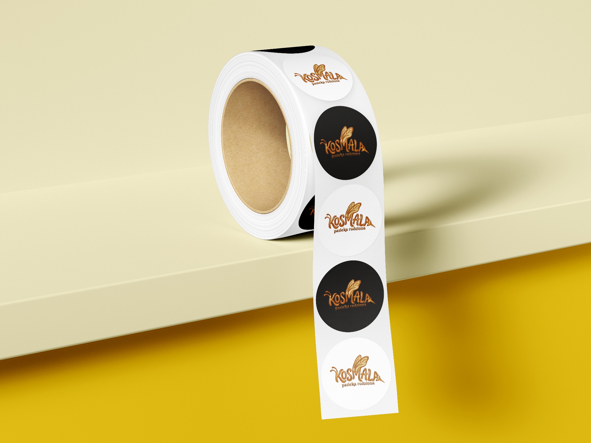 Rolka okrągłych, białych naklejek z brązowym logo 'Kosmala pasieka rodzinna', na żółtym tle. Część naklejek ma czarne tło. Widok z bliska, ostre światło.