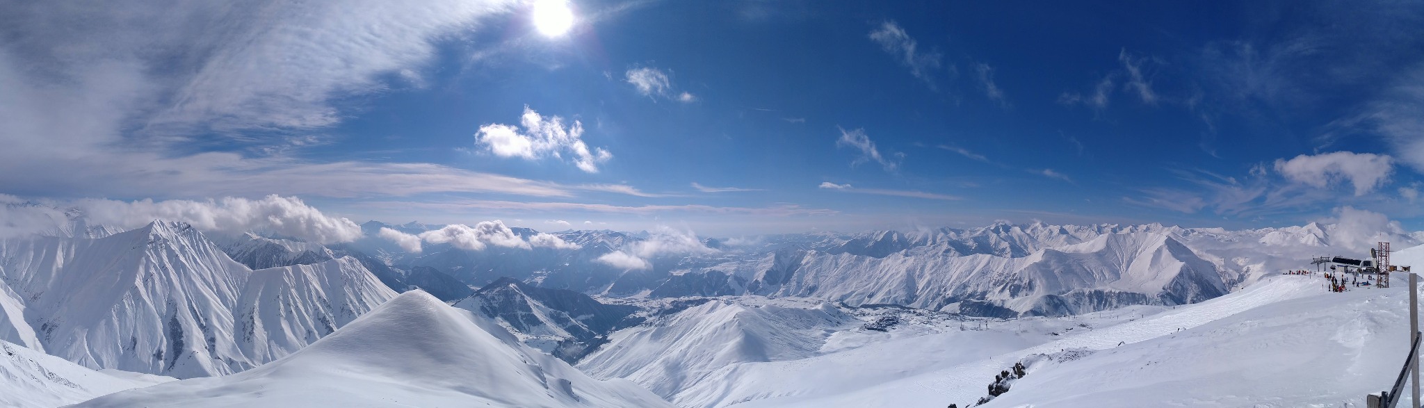 Panoramiczny widok na ośnieżone góry w słoneczny dzień, z widocznymi chmurami i instalacjami wyciągów narciarskich na szczycie.