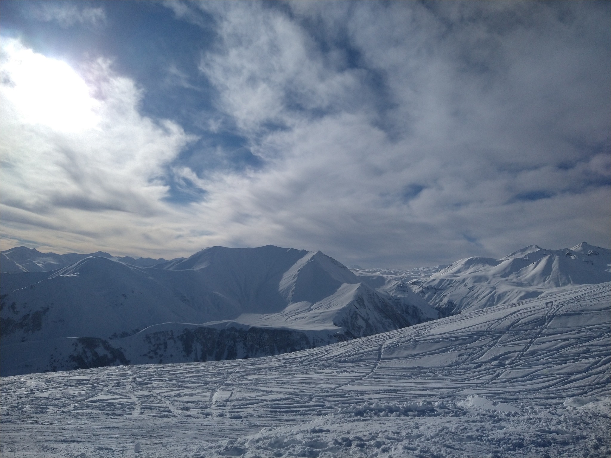 Rozległy, zimowy krajobraz górski z widocznymi śladami nart i snowboardów na śniegu, oświetlony przez słońce przebijające się przez chmury.