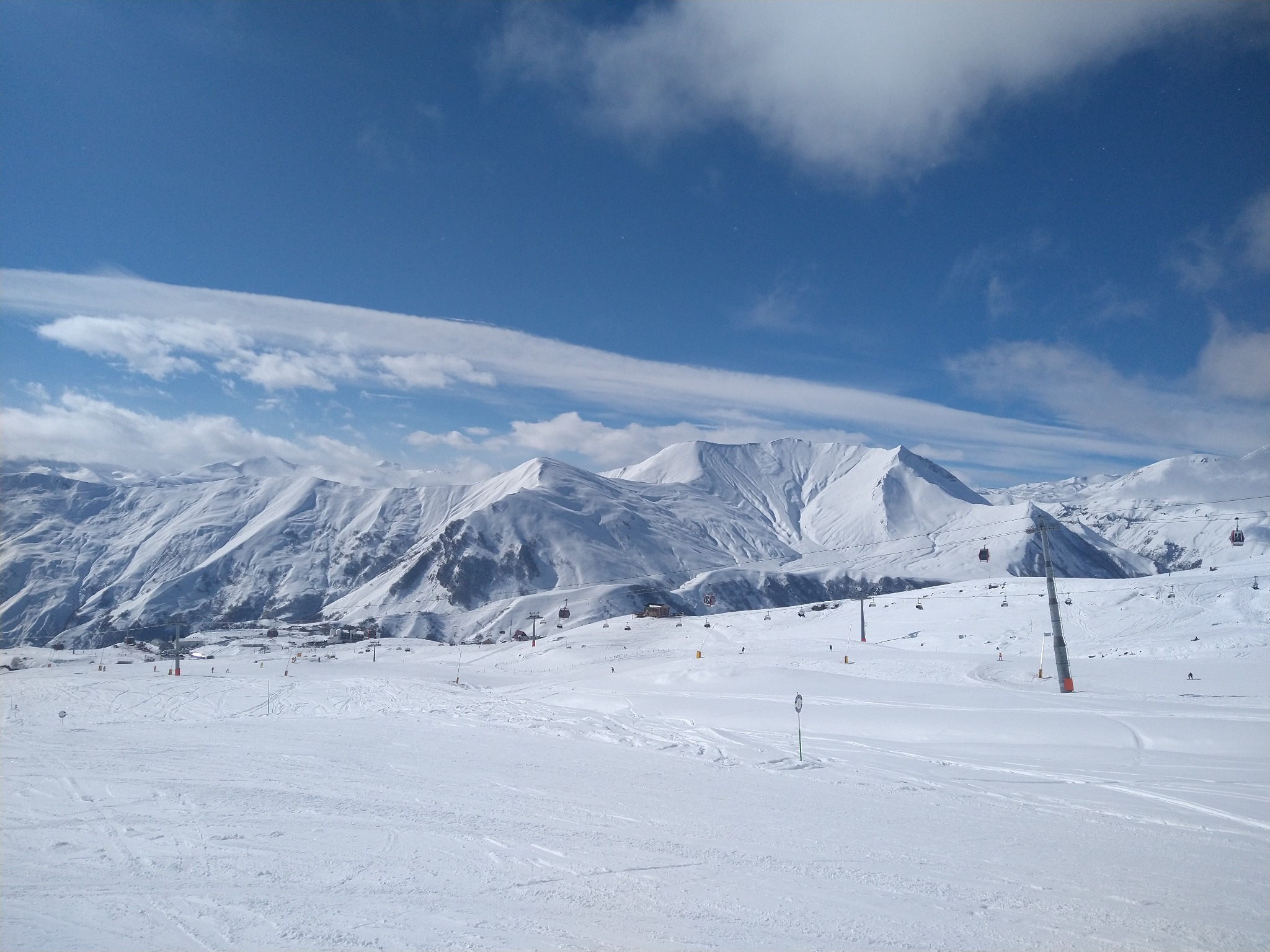 Rozległy, zimowy krajobraz górski z widocznymi trasami narciarskimi, wyciągami krzesełkowymi i gondolami w słoneczny dzień z błękitnym niebem i delikatnymi chmurami.
