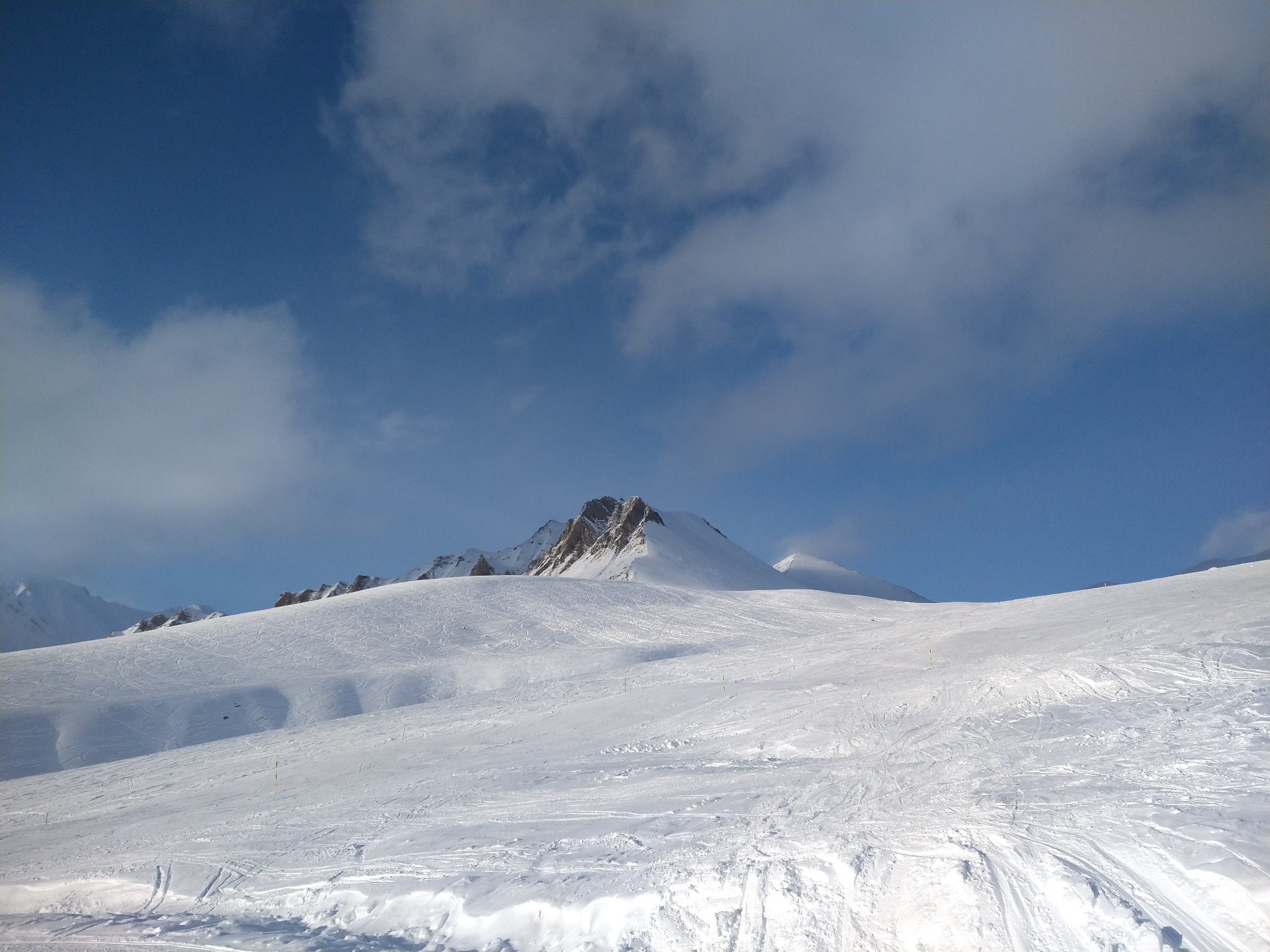 Panoramiczny widok na zaśnieżony stok narciarski z widocznymi śladami po nartach i snowboardach, w tle skaliste szczyty górskie i błękitne niebo z białymi obłokami.