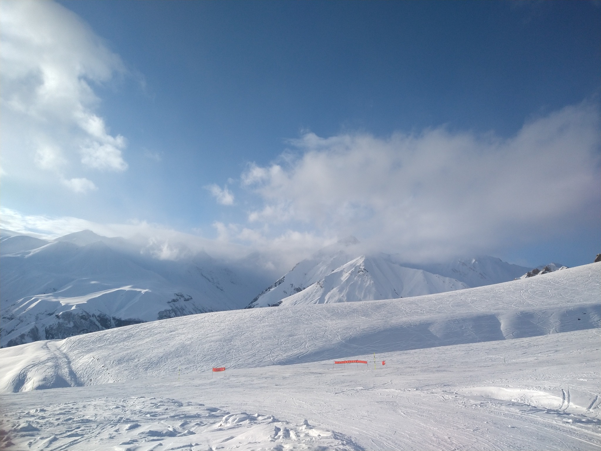 Słoneczny krajobraz górski pokryty śniegiem, z widocznymi śladami nart i pomarańczowymi barierkami zabezpieczającymi trasę.