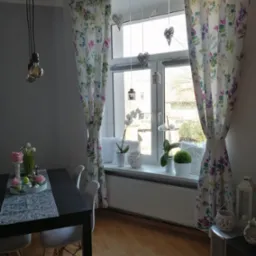 Jasne wnętrze pokoju z oknem udekorowanym wzorzystymi zasłonami w kwiaty, wiszącymi ozdobami w kształcie serc i lampionami; na parapecie białe doniczki z roślinami, a w tle widok na drzewa.