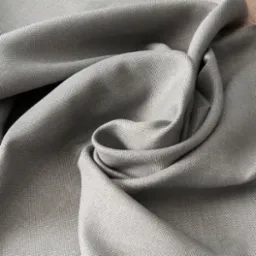Jasnoszara tkanina o wyraźnym splocie, ułożona w miękkie fałdy i delikatne zawirowania, ukazująca strukturę materiału.