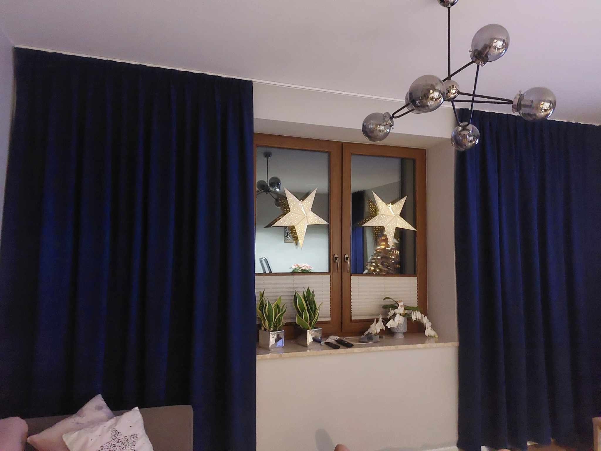 Granatowe zasłony zaciemniające zawieszone przy oknie udekorowanym papierowymi gwiazdami i roślinami doniczkowymi, oświetlone nowoczesną lampą sufitową.