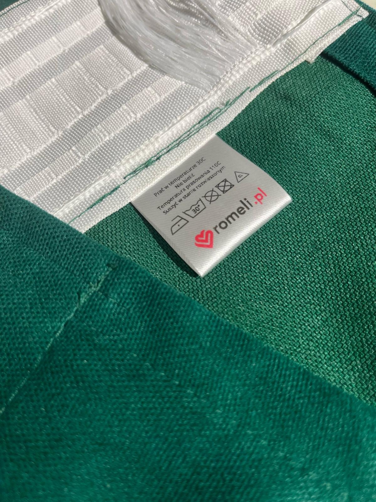Fragment zielonej tkaniny zasłonowej z wszytą białą taśmą marszczącą i metką z instrukcją prania oraz logiem romeli.pl.