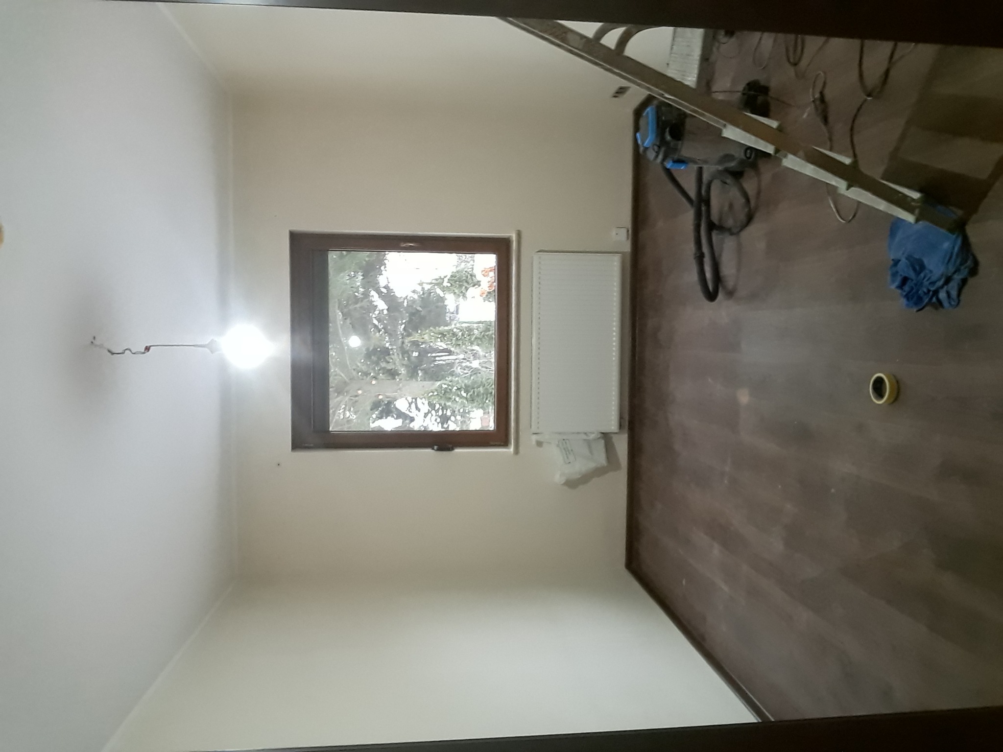 Pokój w trakcie prac wykończeniowych: świeżo pomalowane ściany na biało, częściowo ułożone panele podłogowe, grzejnik zamontowany pod oknem, drabina oparta o ścianę, widoczne narzędzia i materiały...