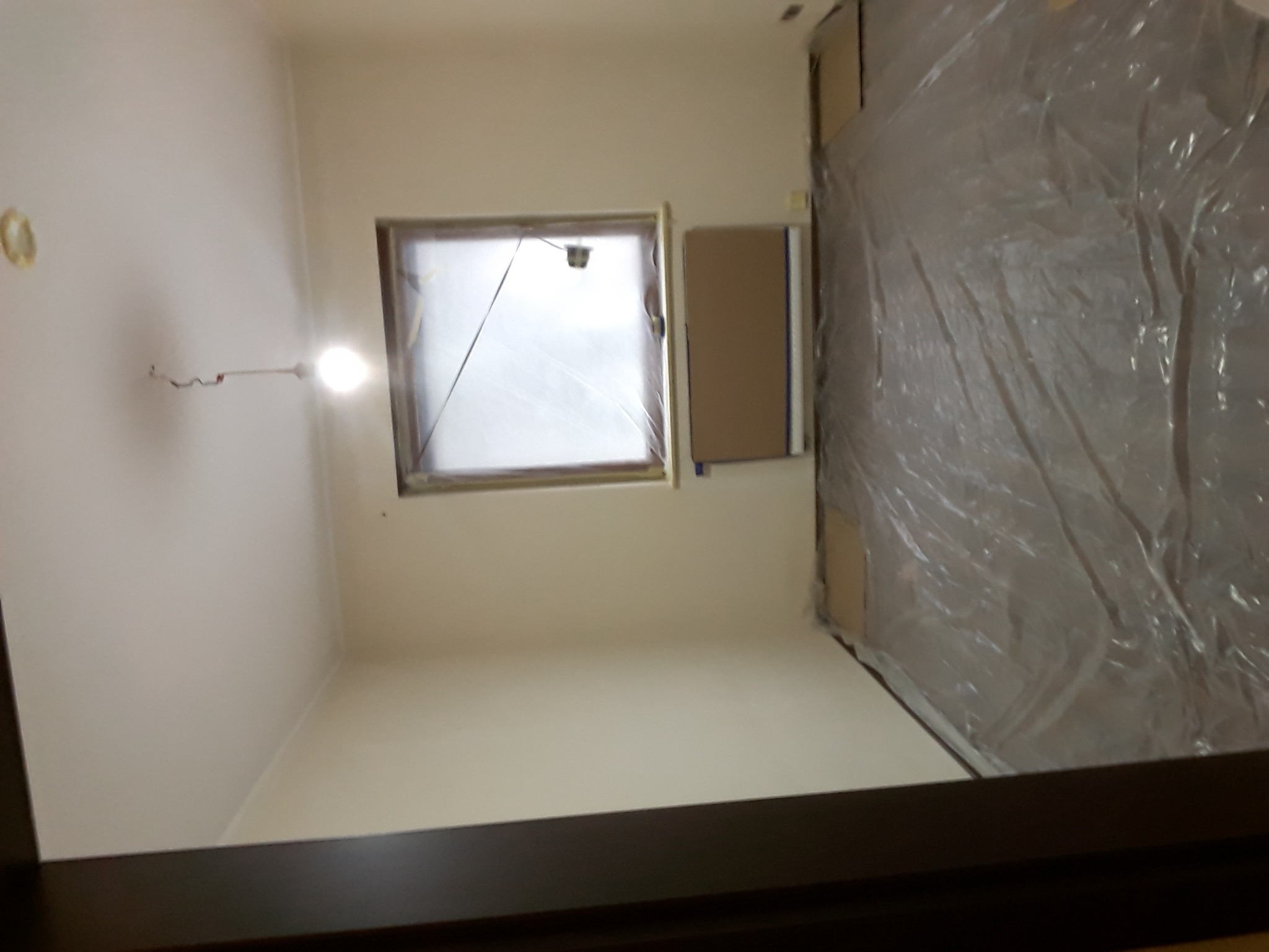 Przygotowane do malowania wnętrze pokoju: okno i fragment ściany zabezpieczone folią malarską, widoczne ślady po gładzi na ścianie.