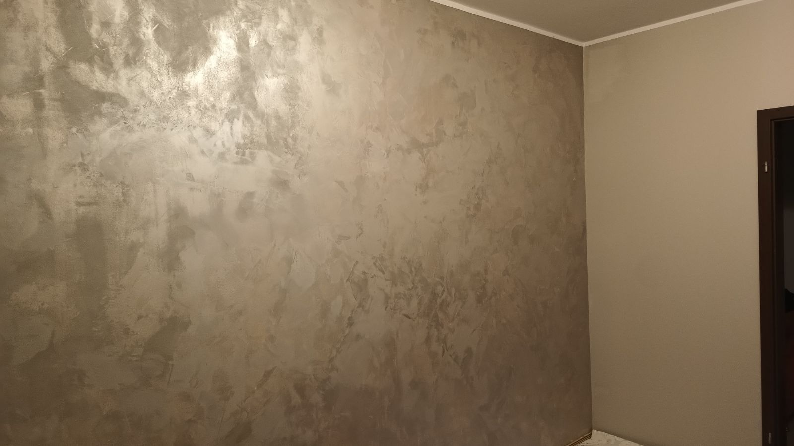 Ściana pokryta dekoracyjną, metaliczną farbą w odcieniach beżu i srebra, widoczna w rogu pokoju obok fragmentu ściany pomalowanej na jednolity, jasny beż i ciemnobrązowej futryny drzwi.