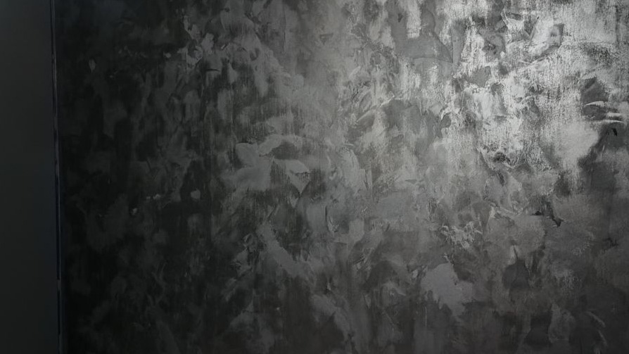Ściana z efektem dekoracyjnym w odcieniach szarości, przypominająca surowy beton lub metal, z widocznymi śladami pędzla i nierównomiernym rozłożeniem farby.