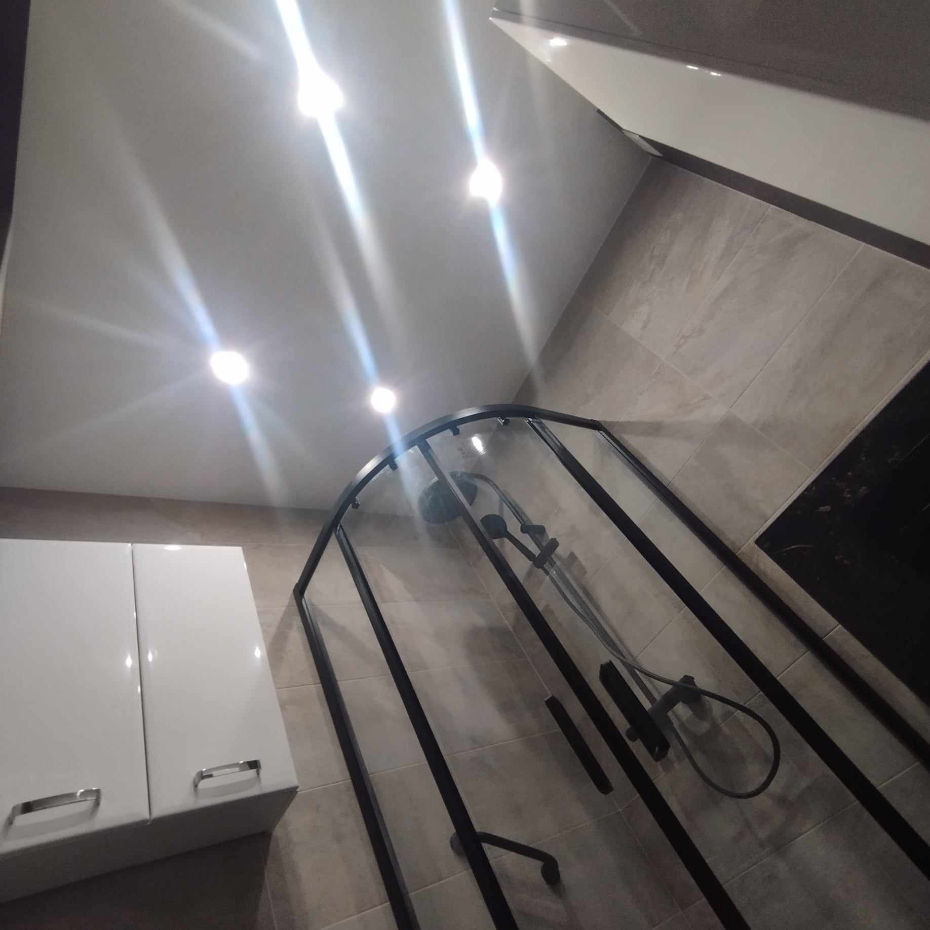 Nowoczesna kabina prysznicowa z czarnymi profilami, zamontowana w łazience z beżowymi płytkami, oświetlona punktowymi lampami sufitowymi, obok biała szafka łazienkowa z chromowanymi uchwytami.