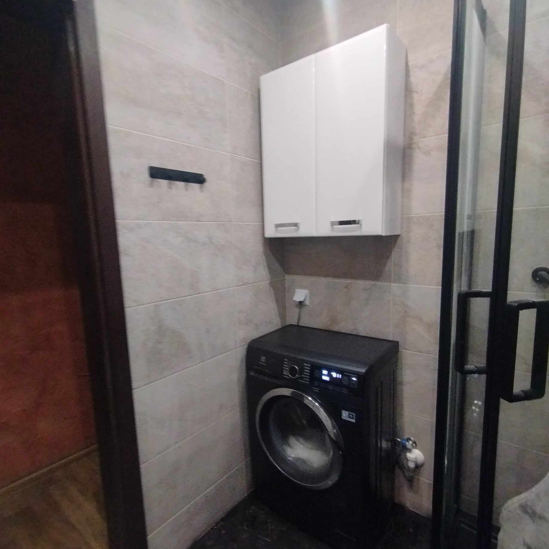 Nowoczesna łazienka z beżowymi płytkami, czarną pralką Electrolux, białą szafką wiszącą i czarną kabiną prysznicową z widoczną armaturą.