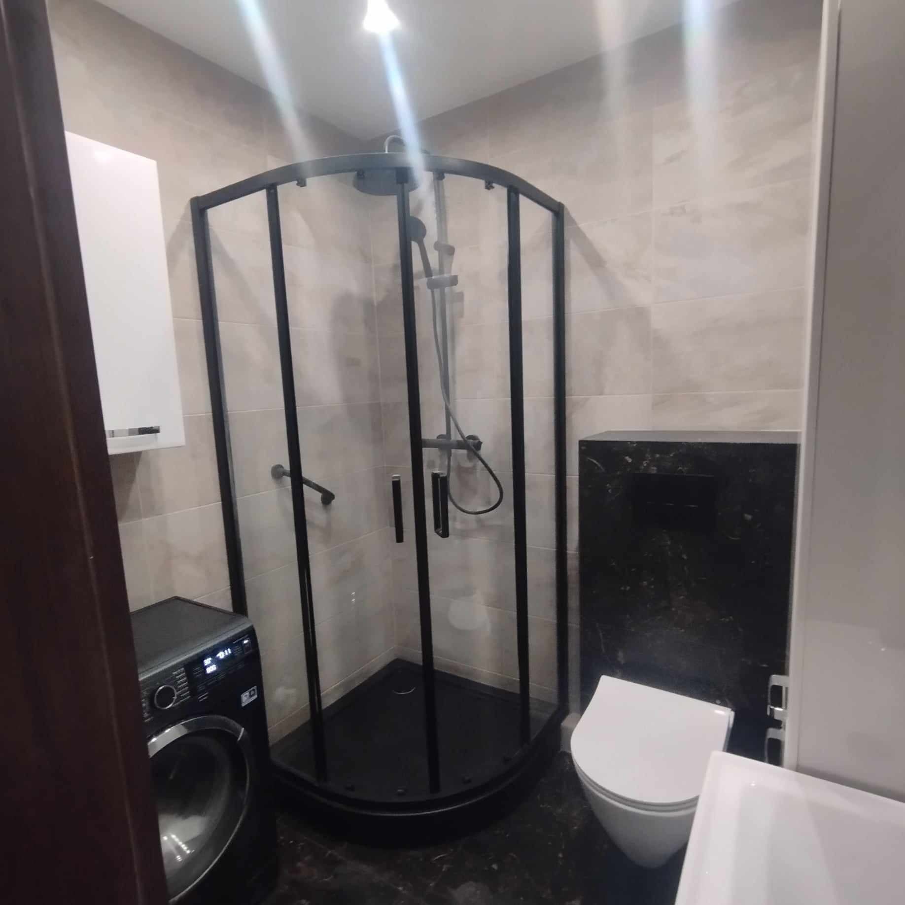 Nowoczesna łazienka z czarną kabiną prysznicową narożną, umywalką, toaletą i pralką w odcieniach beżu i czerni.