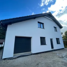 Nowoczesny, dwukondygnacyjny dom z białymi ścianami, czarnym dachem i stolarką okienną, widziany z poziomu gruntu, z niezagospodarowanym terenem wokół.