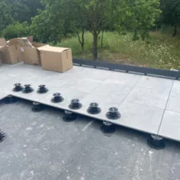 Montaż płytek na regulowanych wspornikach na dachu, widoczne kartony z płytkami i zieleń w tle.