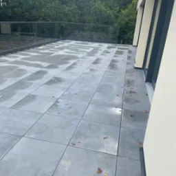 Nowoczesny taras z szarych, kwadratowych płyt, zabezpieczony szklaną balustradą, z widocznymi liśćmi na powierzchni i fragmentem elewacji budynku.