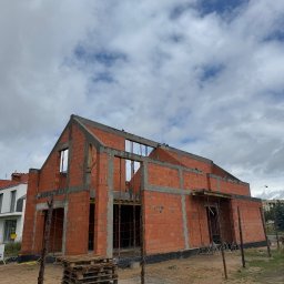Zakład Ogólnobudowlany Marek Masłek - Dom jednorodzinny w trakcie budowy, z murami z czerwonych pustaków ceramicznych i widocznymi betonowymi elementami konstrukcyjnymi, pod zachmurzonym niebem.