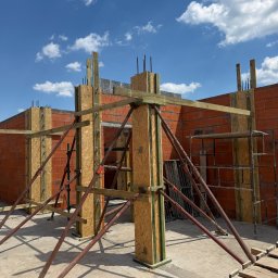 Zakład Ogólnobudowlany Marek Masłek - Szalunek z płyt OSB i drewnianych belek pod betonowe słupy i ściany, stabilizowany podporami, na budowie domu z czerwonej cegły, pod błękitnym niebem.