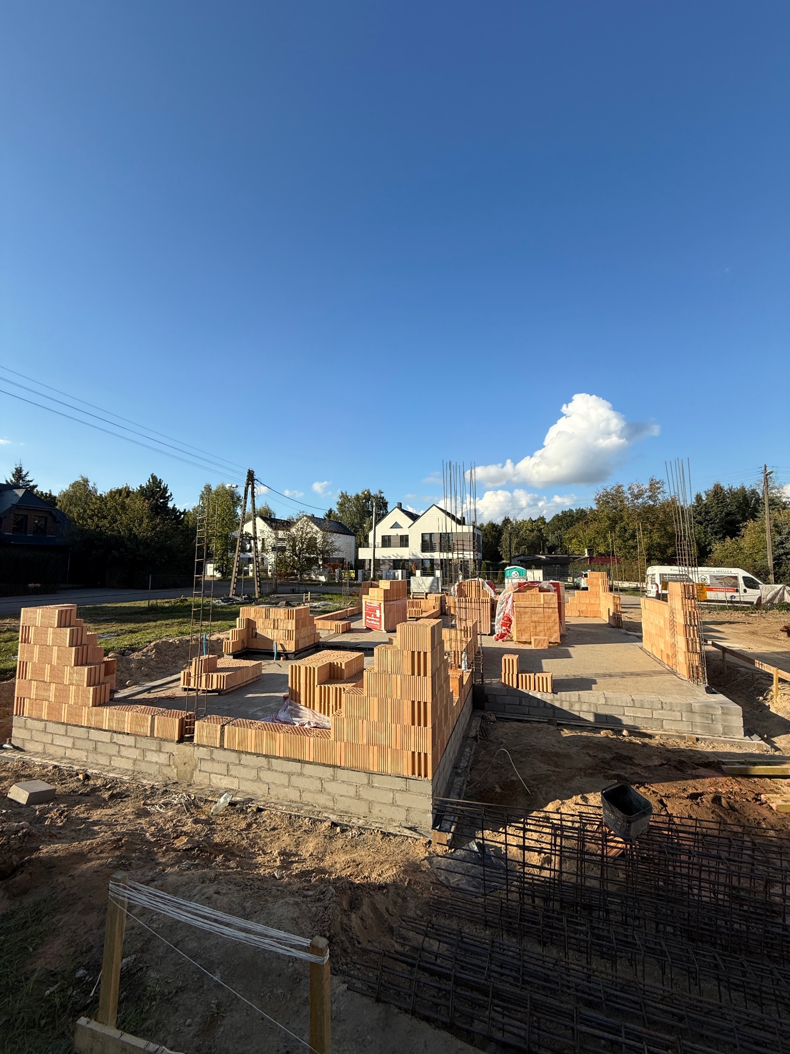 Wczesny etap budowy domu z cegły ceramicznej: fundamenty, pierwsze warstwy ścian, wystające pręty zbrojeniowe. W tle domy jednorodzinne i błękitne niebo.