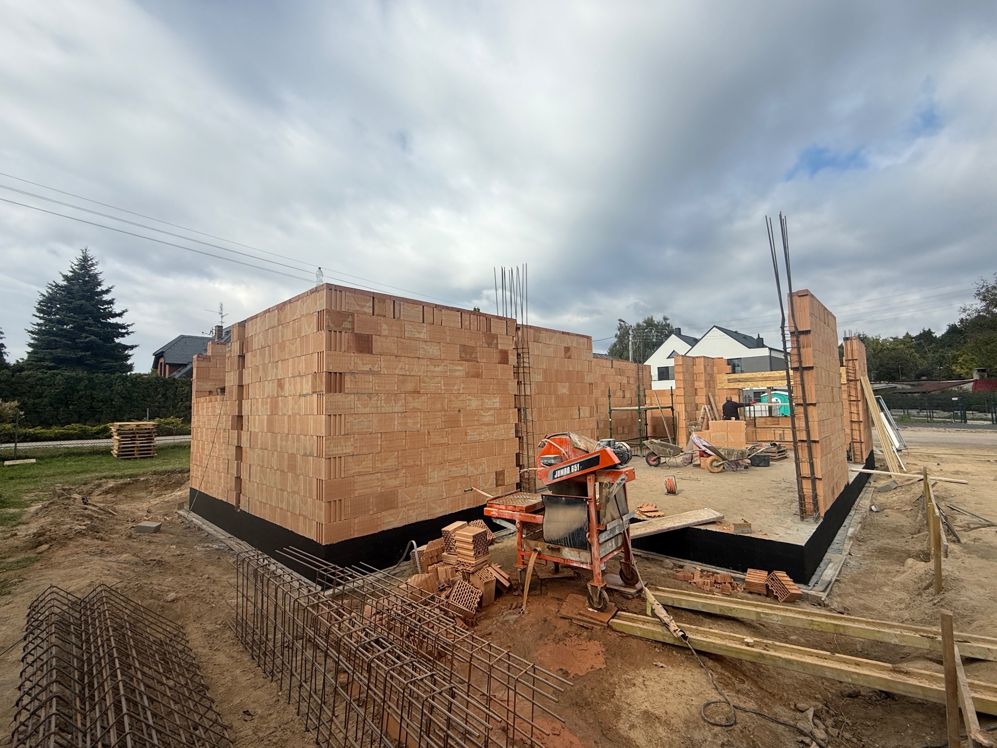 Budowa domu z czerwonej cegły w Czarnkowie, widoczne zbrojenia i narzędzia. Ściany wzniesione do pewnej wysokości, przygotowane do dalszych prac.