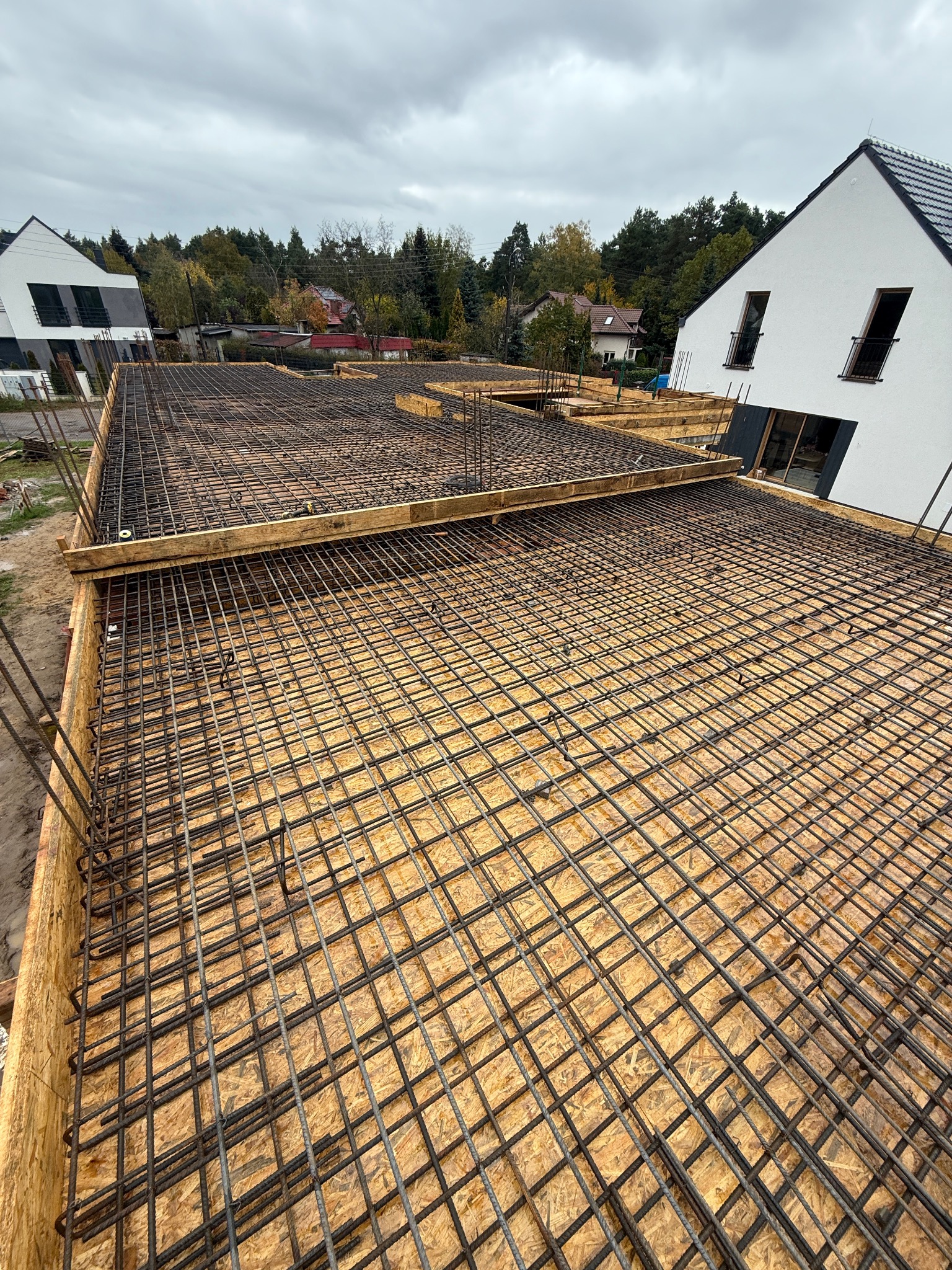 Zbrojenie płyty fundamentowej domu jednorodzinnego z widoczną siatką stalową i deskowaniem, w tle zabudowa jednorodzinna i pochmurne niebo.