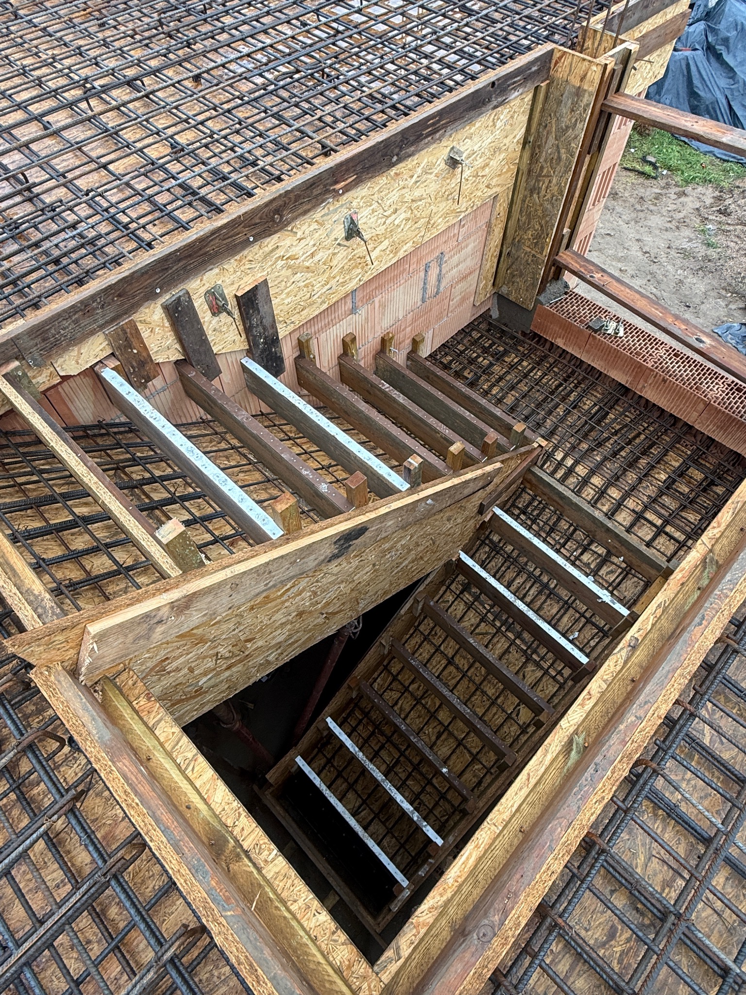 Zbrojenie schodów z drewnianym szalunkiem i ceglanymi elementami, widok z góry. Metalowa siatka zbrojeniowa na górze i schodach. Etap budowy.