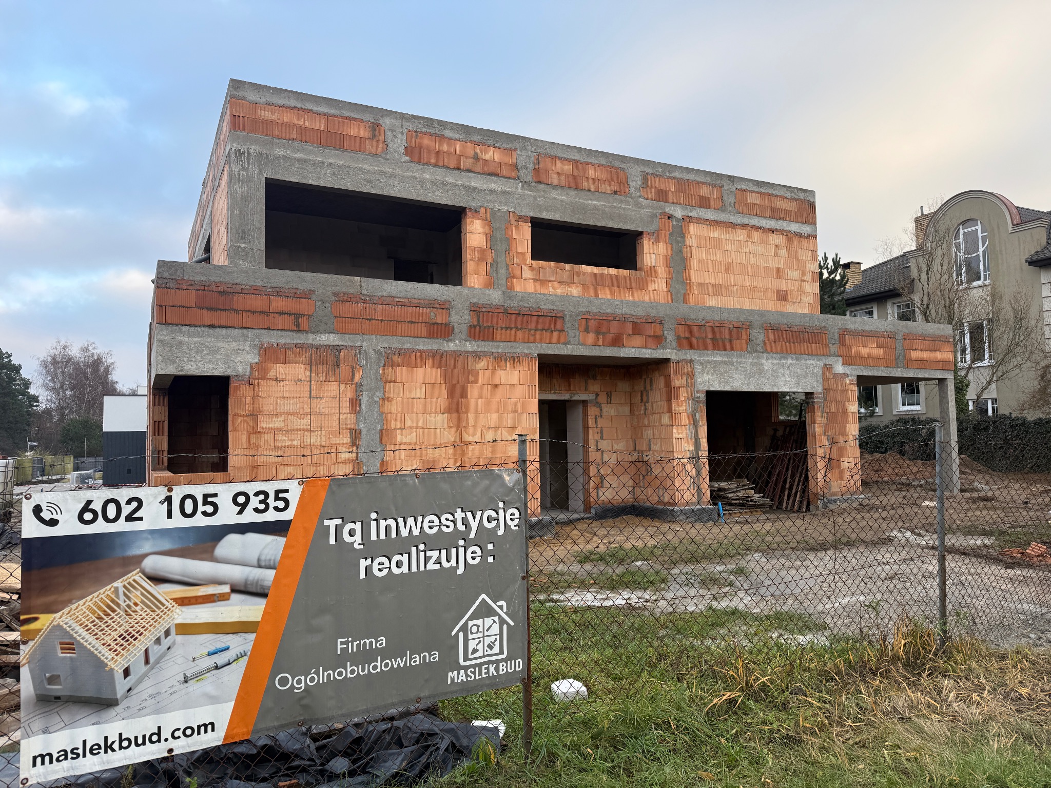 Surowy stan domu z czerwonej cegły, widoczny przez siatkę ogrodzeniową z tablicą reklamową firmy budowlanej Maslek Bud. W tle dom z oknem w kształcie łuku.
