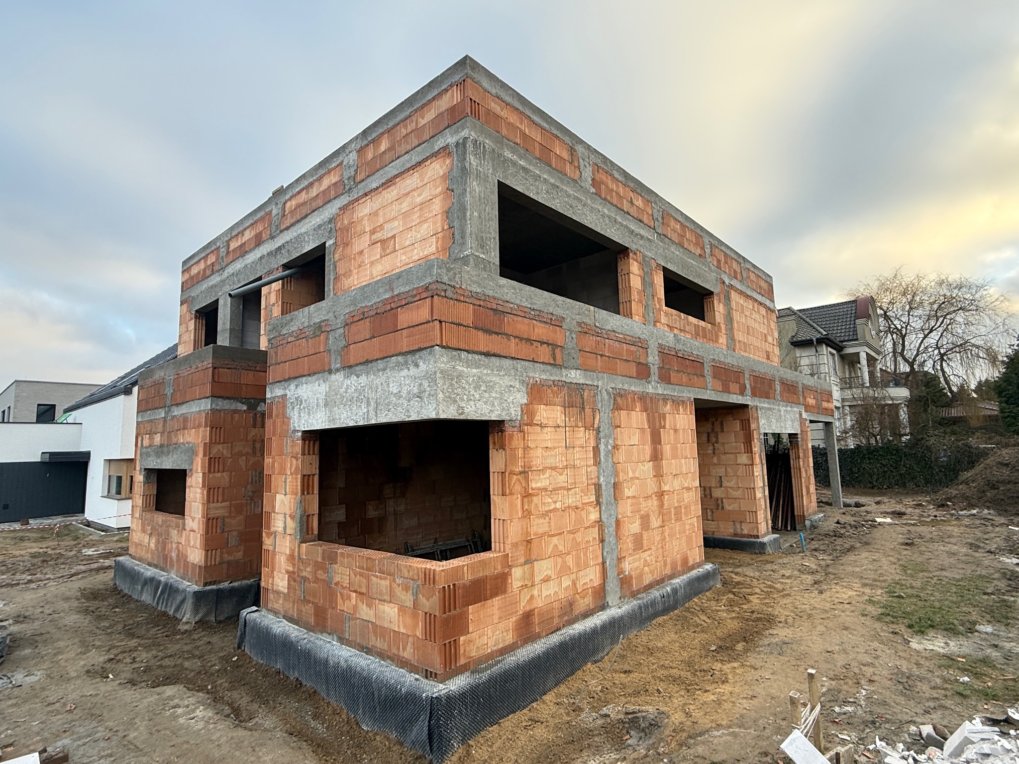 Surowy stan dwukondygnacyjnego domu z czerwonej cegły i betonu, widok z zewnątrz, w tle inne budynki i pochmurne niebo. Fundament zabezpieczony czarną folią.