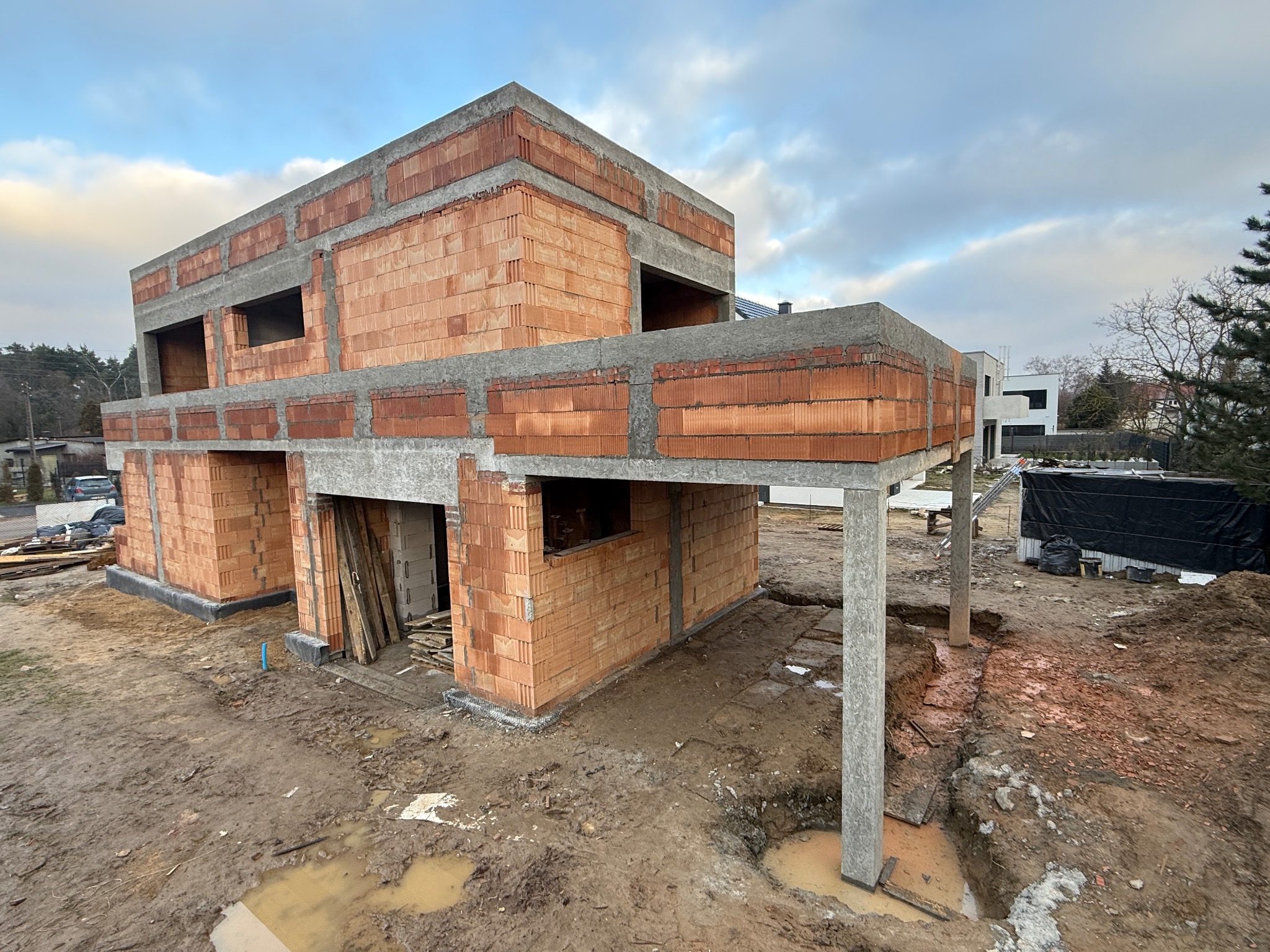 Surowy stan dwukondygnacyjnego domu z czerwonej cegły, z widocznymi betonowymi nadprożami i słupami, na tle pochmurnego nieba i sąsiednich budynków.