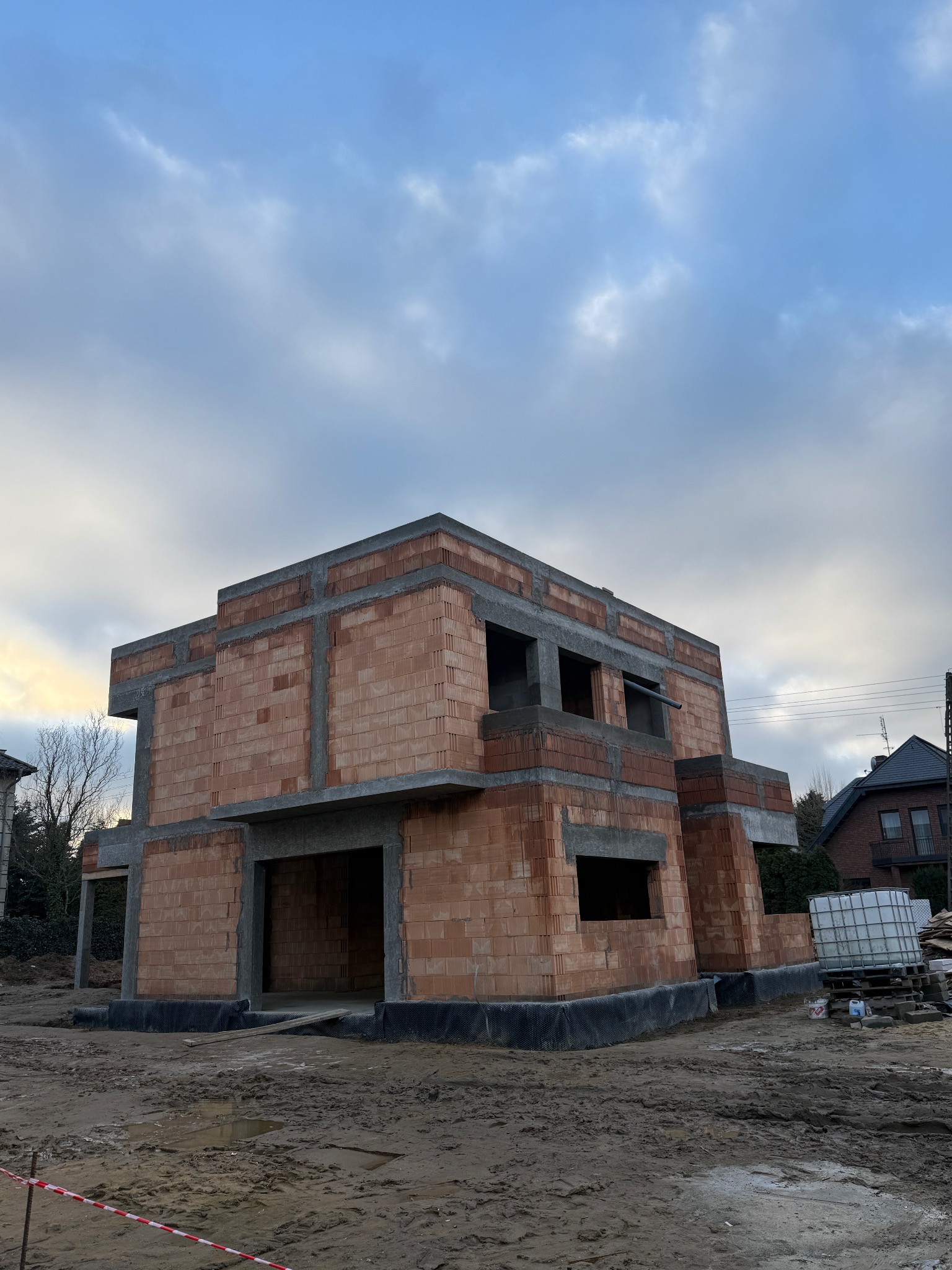 Surowy stan dwukondygnacyjnego domu z czerwonej cegły, z widocznymi betonowymi wzmocnieniami, na tle pochmurnego nieba i sąsiednich budynków.